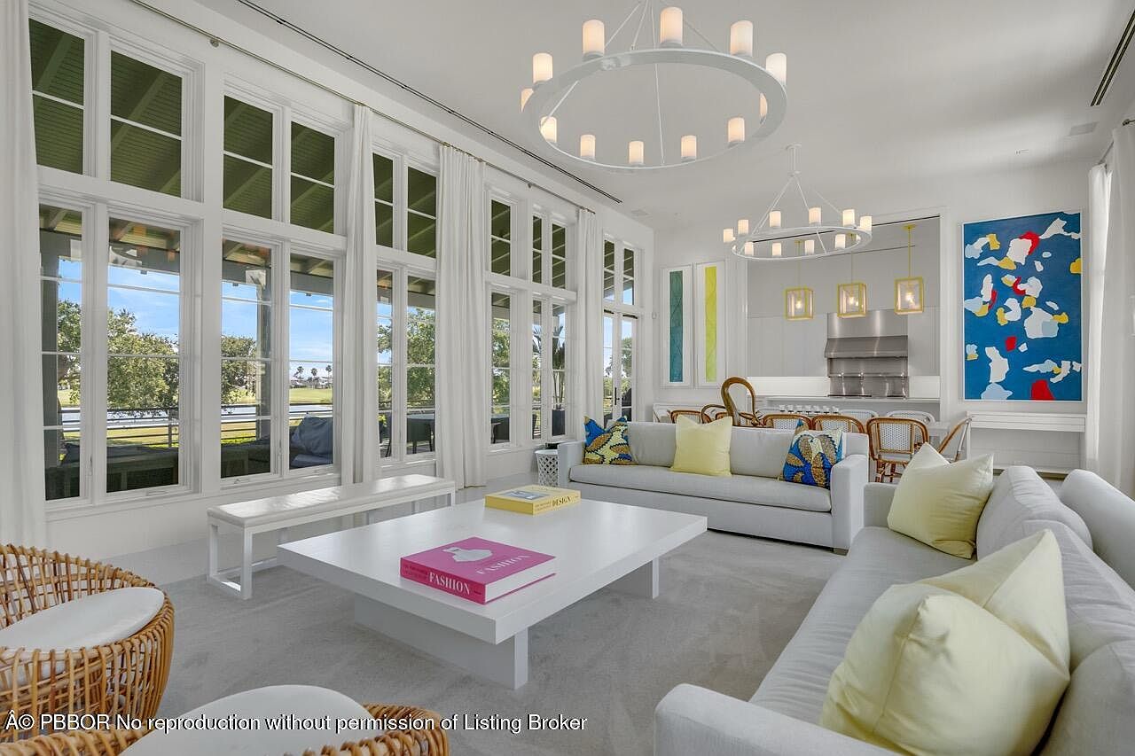 This is a bright and airy living room featuring large windows that offer an expansive view. The room is furnished with white sofas, a white coffee table with fashion books, and wicker chairs, creating a clean and modern aesthetic. Two modern chandeliers hang from the ceiling, adding a touch of elegance.