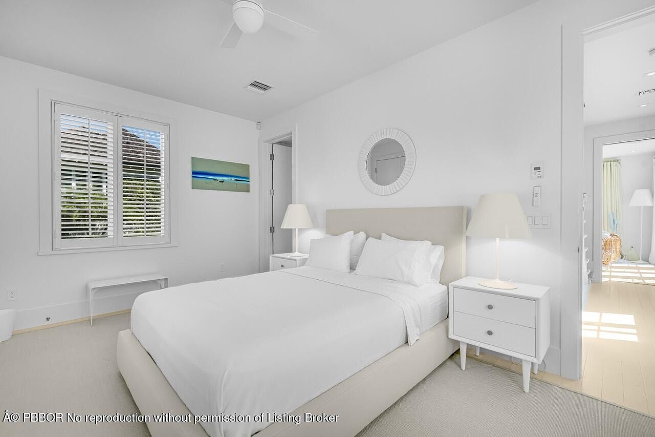 This is a bright and airy primary bedroom featuring a neutral color palette. The room is furnished with a large bed with white linens, two white nightstands with lamps, and a round mirror above the bed. Natural light streams in through the window, creating a serene and inviting atmosphere.