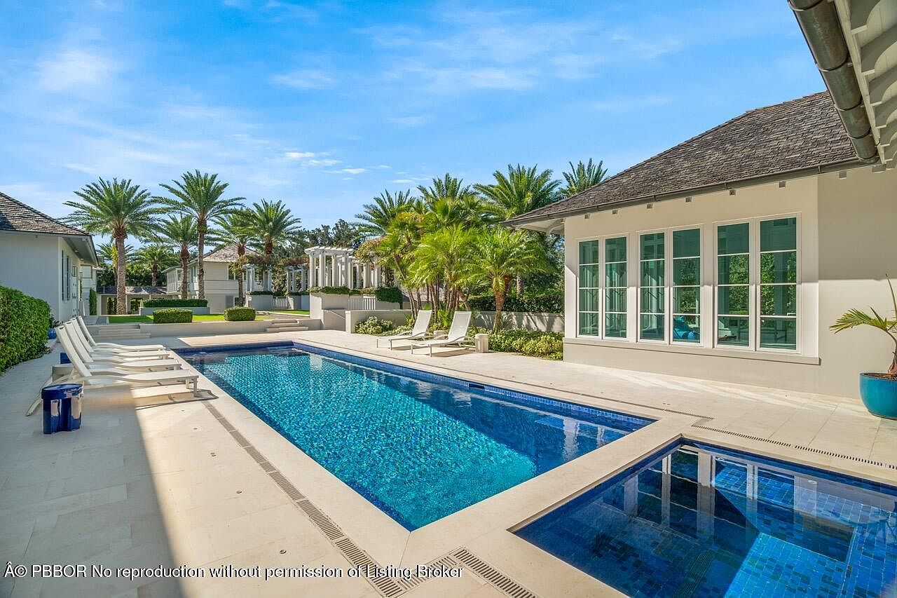 This image showcases a luxurious pool area with a rectangular pool featuring blue mosaic tiles, complemented by a smaller spa pool. The surrounding patio is made of light-colored stone, and lounge chairs are arranged for relaxation. Palm trees and manicured landscaping add to the tropical ambiance, while the architecture of the adjacent building features large windows and a neutral color palette.
