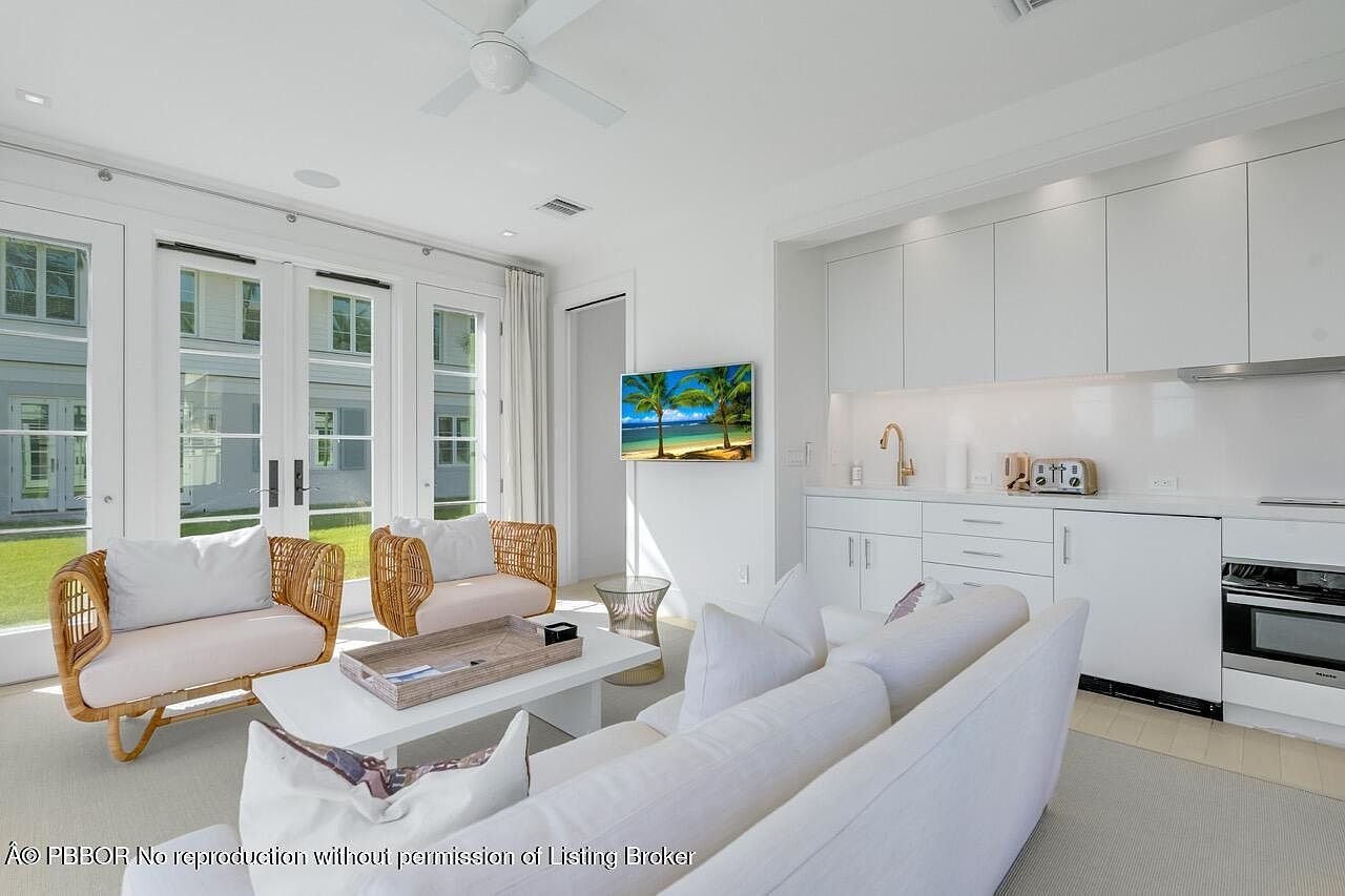 This is a bright and airy living room featuring a white sofa, two wicker chairs with light cushions, and a white coffee table. The room has a modern, clean aesthetic with white walls and cabinetry, and a flat-screen TV mounted on the wall. Large windows and glass doors provide ample natural light and a view of the outdoors.