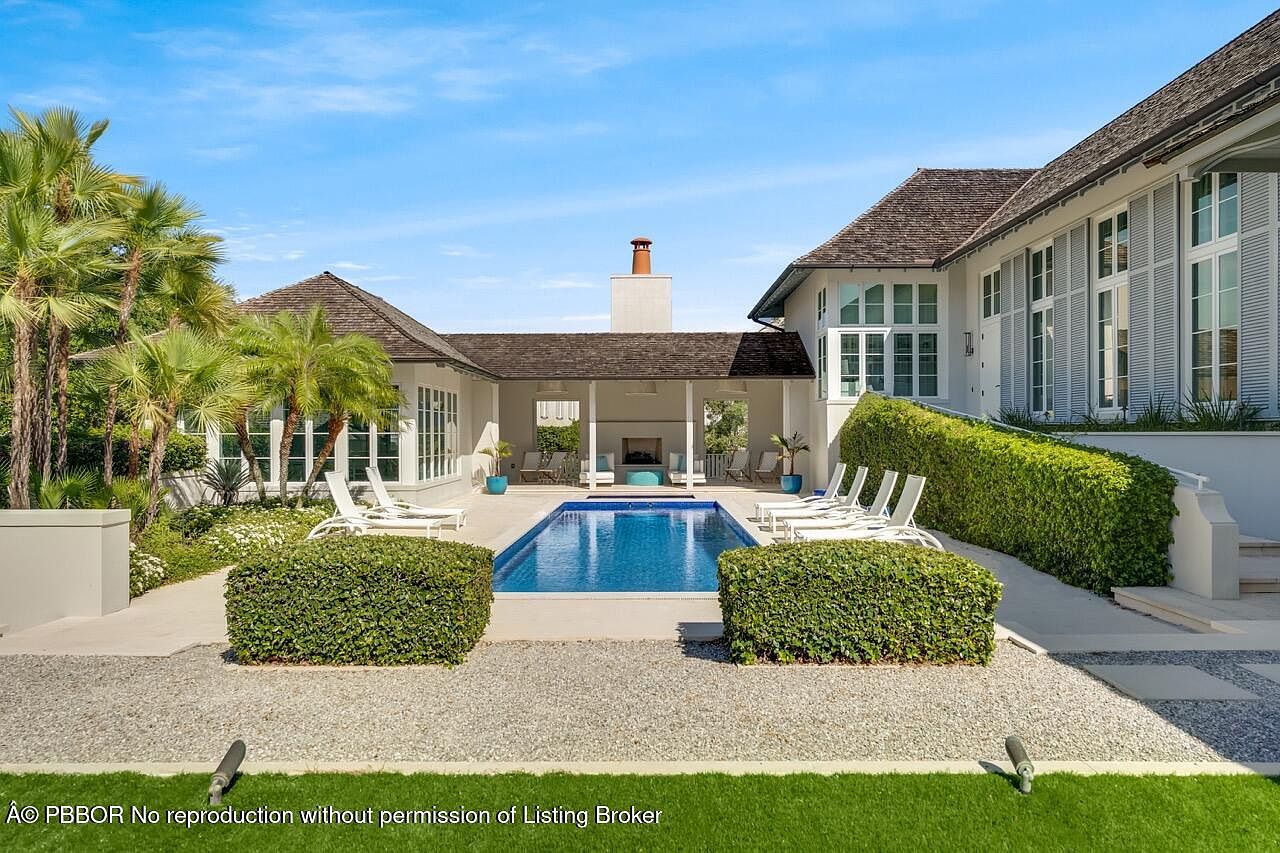 This image showcases a luxurious backyard pool area. The rectangular pool is surrounded by manicured greenery, including trimmed hedges and palm trees. Lounge chairs are neatly arranged around the pool, and the architecture of the house features white walls, dark roofing, and a covered patio area with an outdoor fireplace, creating an inviting and upscale outdoor living space.