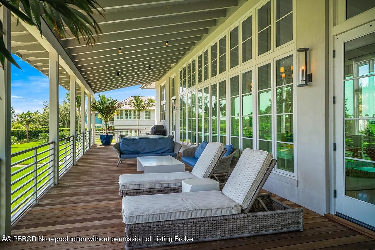 This image showcases a luxurious outdoor patio or deck area. The space is furnished with comfortable lounge chairs and a sofa, suggesting a relaxing atmosphere. The deck is made of wood and is surrounded by a railing, offering views of the well-manicured lawn and the house in the background. Large windows and a door provide access to the interior, and a wall sconce adds a touch of elegance.