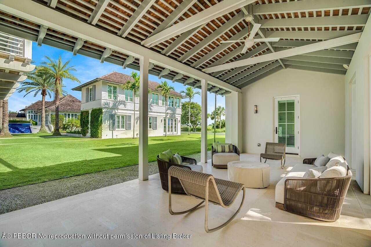 This image showcases a luxurious outdoor patio area, featuring comfortable wicker seating arrangements under a covered structure with exposed beams. The patio overlooks a sprawling green lawn and a well-maintained house, creating a serene and inviting atmosphere. The perspective is from the patio looking out towards the lawn and house.