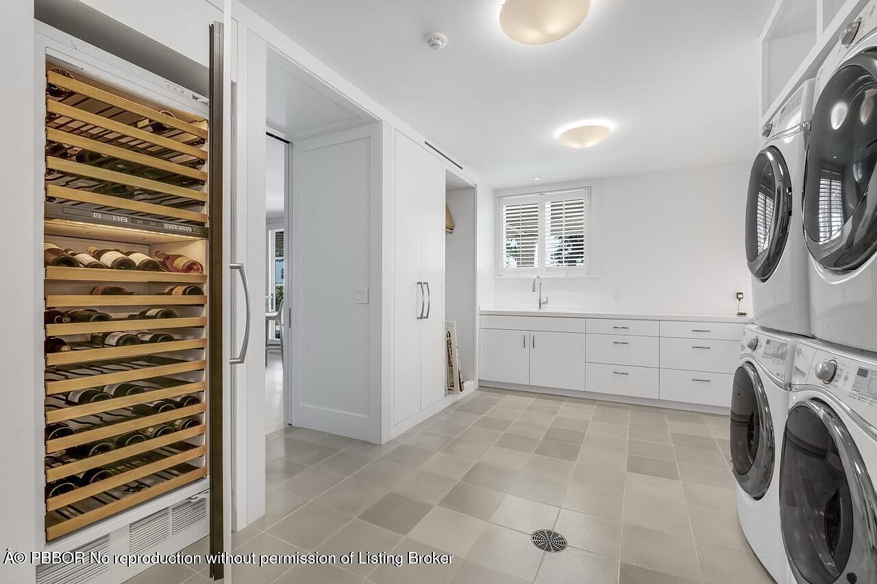 This is a well-organized laundry room featuring a wine refrigerator, stacked washer and dryer, and ample counter space with built-in cabinets. The room is brightly lit with recessed lighting and a window with blinds, and the neutral tile flooring adds a clean, modern touch. The perspective is from the doorway, capturing the full scope of the room's functionality and design.