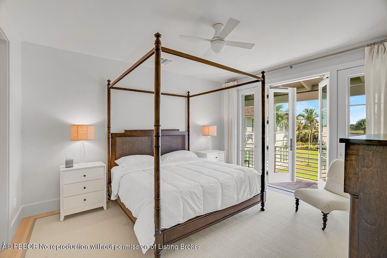 This is a bright and airy primary bedroom featuring a four-poster bed with white linens, complemented by matching white nightstands and lamps. French doors open to a balcony, offering a view of the outdoors and filling the room with natural light. The room is decorated in a neutral color palette, creating a serene and inviting atmosphere.