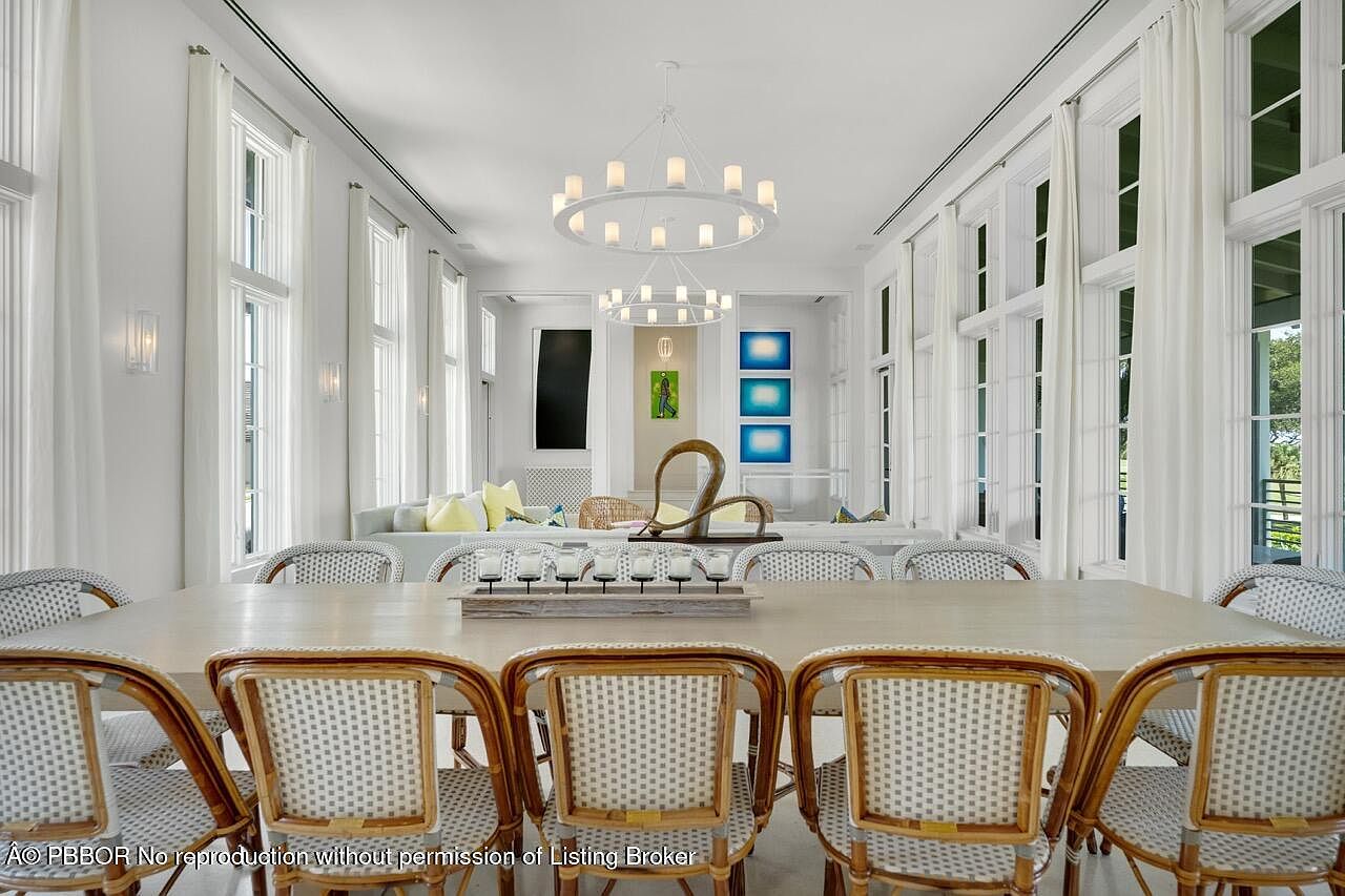 This is an interior shot of a bright and airy dining room. The room features a long wooden dining table surrounded by wicker chairs, illuminated by two modern circular chandeliers. Large windows with white curtains line the walls, providing ample natural light and creating a sophisticated and inviting atmosphere.