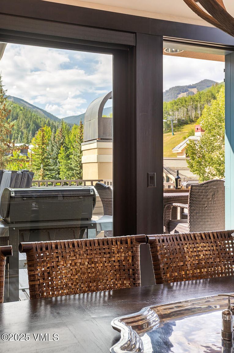 This image showcases a patio or balcony area with a view of a mountain landscape. The space includes outdoor furniture, such as wicker chairs and a dining table, suggesting an area for relaxation and dining. A grill is also visible, indicating the potential for outdoor cooking and entertaining.