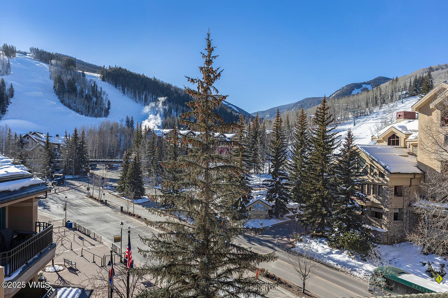 This image showcases a scenic view of a property in a snowy, mountainous area. The foreground features a well-maintained street and sidewalk, leading to buildings with stone and wood exteriors. In the background, a ski slope is visible, suggesting a location ideal for winter sports enthusiasts. The overall impression is one of a luxurious mountain resort.