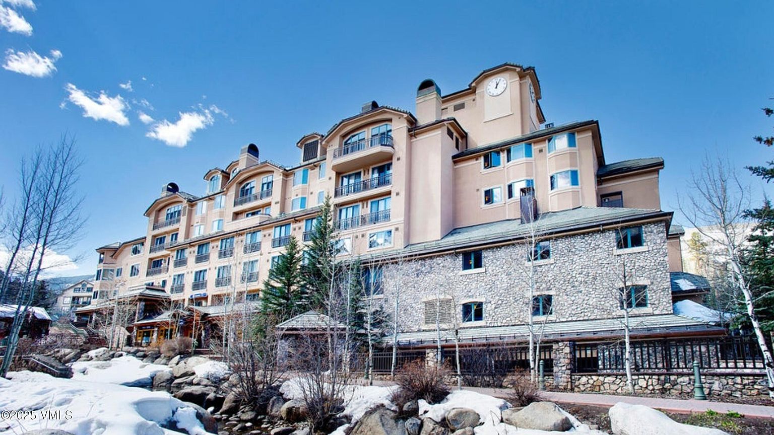 This is a front exterior view of a multi-story building, possibly a condominium or resort, showcasing a blend of stone and stucco facade. The architecture features multiple balconies and a prominent clock tower. The surrounding landscape includes snow-covered areas, rocks, and bare trees, suggesting a mountain or ski resort location.