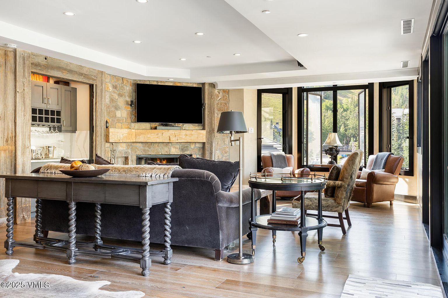 This is an interior shot of a living room featuring a stone fireplace with a mounted television above it. The room is furnished with a dark gray sofa, a wooden console table, and a round side table with books. Large windows provide natural light and a view of the outdoors, creating a cozy and inviting atmosphere.