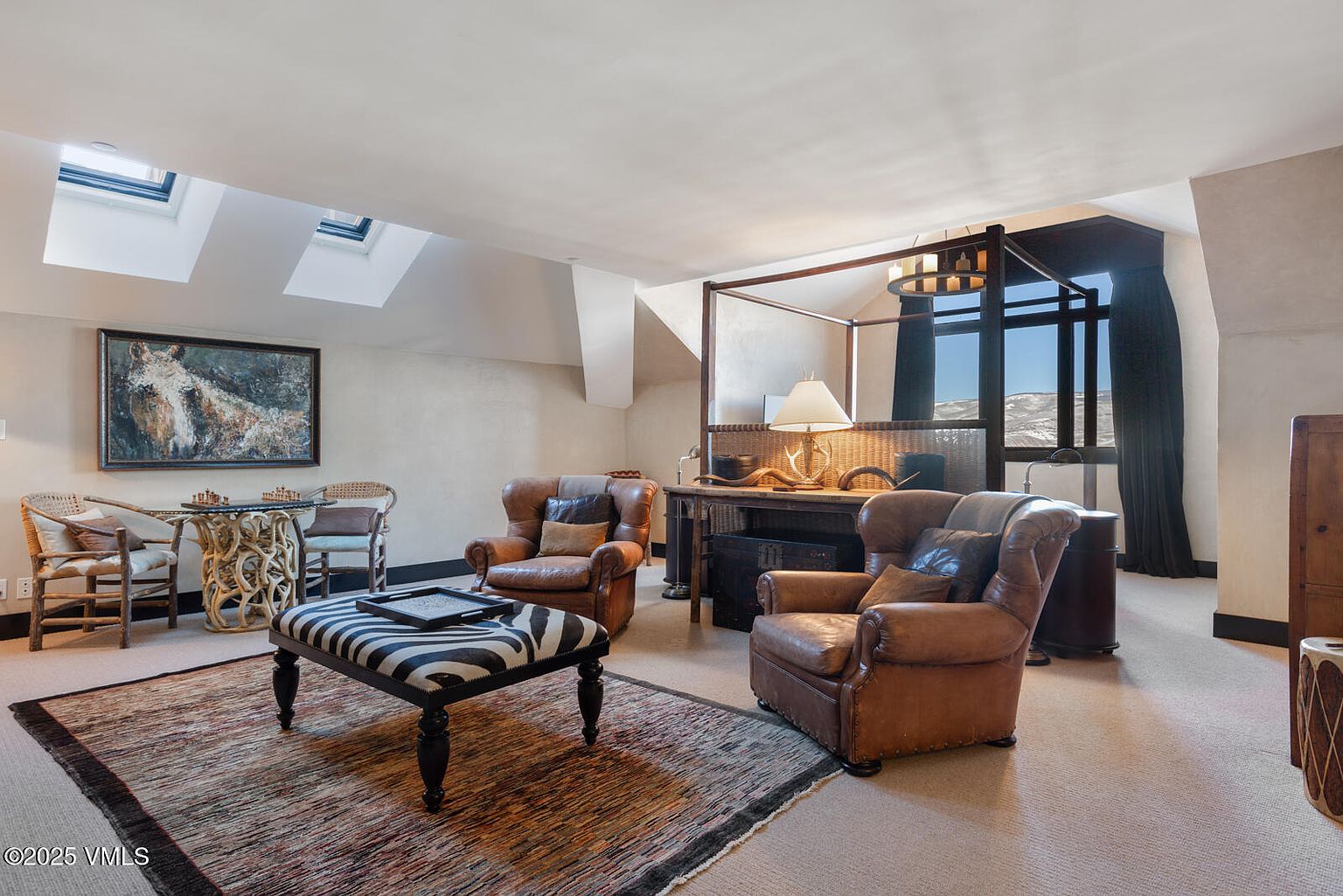 This is an interior shot of a cozy living room featuring two leather armchairs, a zebra-striped ottoman, and a unique glass-topped table with wicker chairs. The room is well-lit by skylights and a window offering a view of a snowy landscape, complemented by a four-poster structure and a desk with a lamp. The overall style is rustic and inviting, perfect for a relaxing retreat.