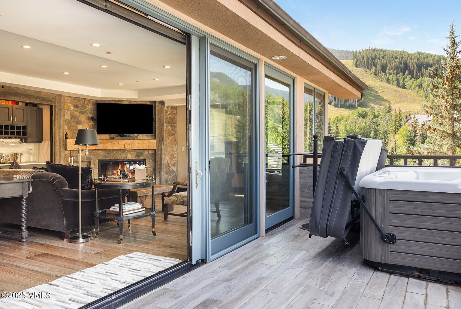 This image showcases a luxurious patio or balcony area, seamlessly blending indoor and outdoor living. Sliding glass doors open to reveal a stylish living room with a stone fireplace and comfortable seating. The outdoor space features a hot tub, offering a relaxing retreat with scenic mountain views in the background.