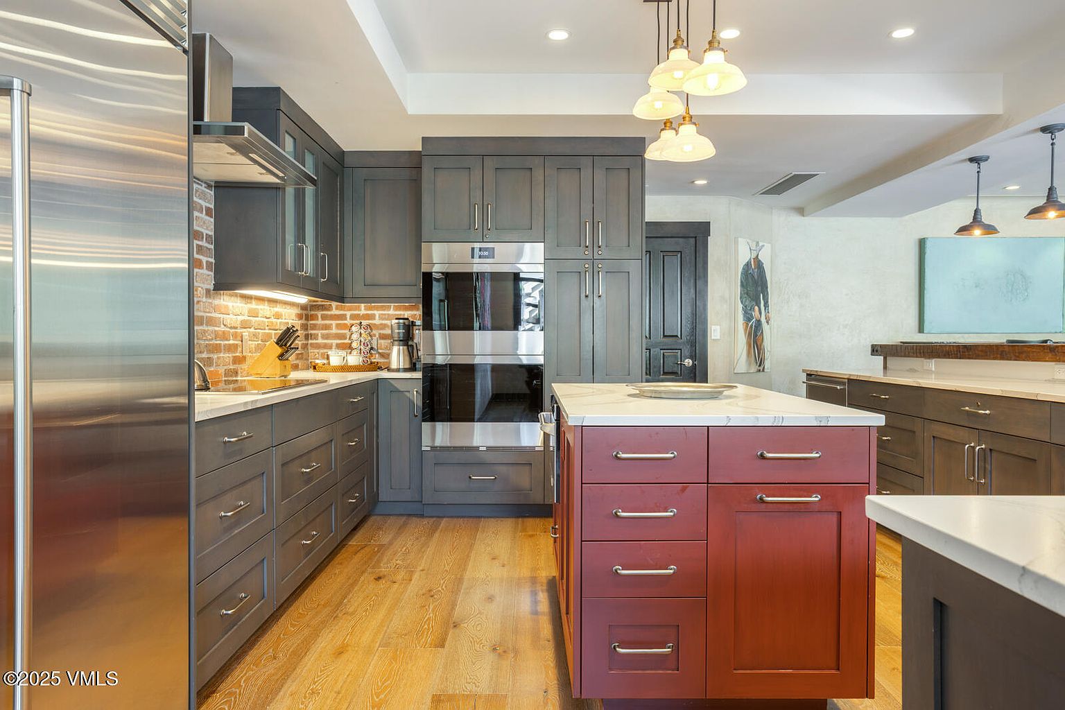 This is a well-appointed kitchen featuring gray cabinetry, stainless steel appliances, and a contrasting red island with a white countertop. The kitchen also has a brick backsplash and hardwood floors. The perspective is from the front, showcasing the layout and design elements of the space.