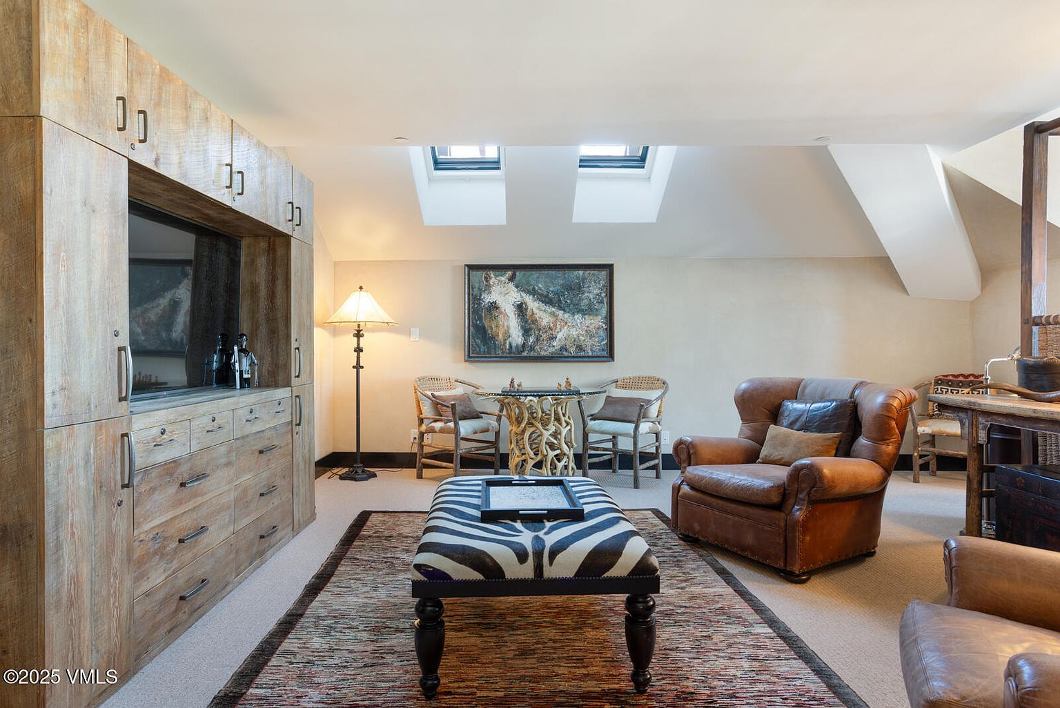 This inviting living room features a built-in wooden cabinet with a television, a zebra-striped ottoman, and comfortable leather armchairs. A decorative table with wicker chairs sits beneath a framed painting, and natural light streams in through skylights. The room exudes a warm and rustic elegance.