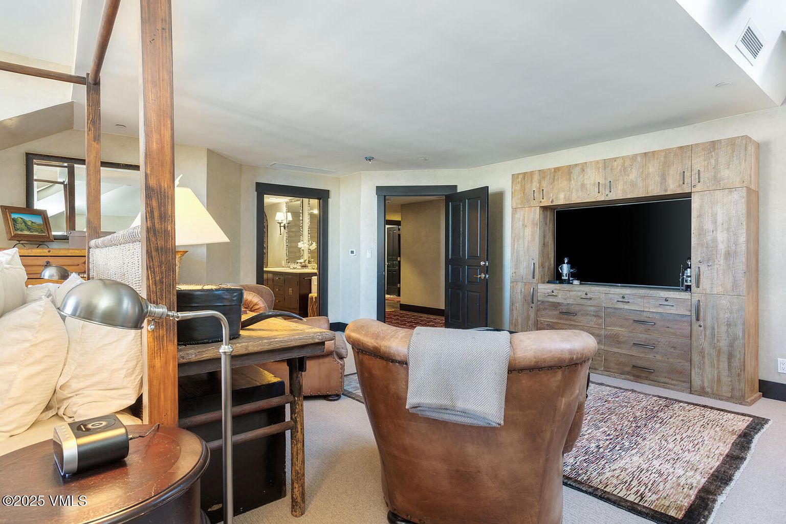 This primary bedroom features a rustic yet luxurious design with a four-poster bed, a wooden entertainment center housing a large television, and a comfortable leather armchair. The room includes access to a bathroom and another room, enhancing its functionality and flow. The overall aesthetic is warm and inviting, blending natural materials with modern amenities.