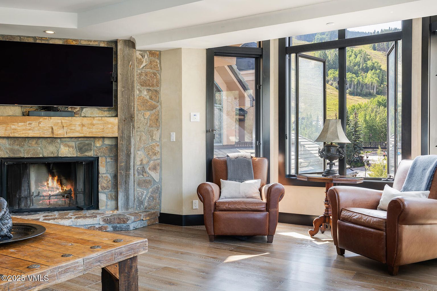 This inviting living room features a stone fireplace with a mounted television above, creating a cozy focal point. Two leather armchairs are positioned near a large window offering scenic views of the outdoors. The hardwood flooring and natural light enhance the room's warm and comfortable atmosphere.