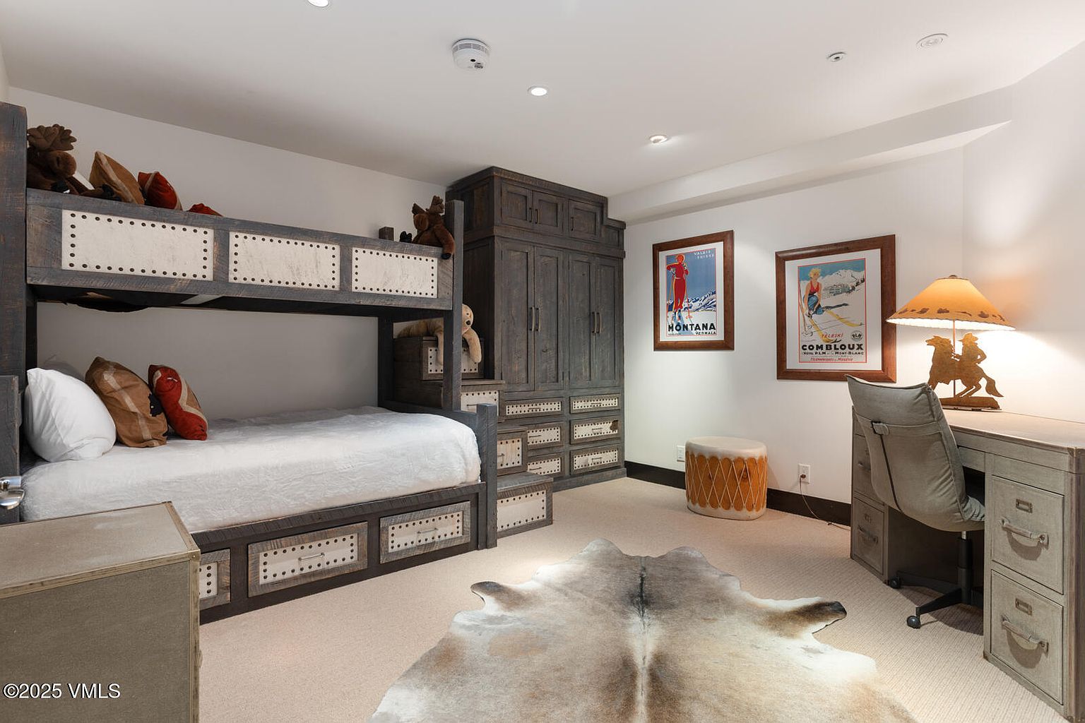This is a cozy guest bedroom featuring a custom-built bunk bed with storage drawers underneath, complemented by a matching wardrobe and desk. The room is decorated with rustic accents, including framed vintage ski posters and a cowhide rug, creating a warm and inviting atmosphere. The perspective is from a medium shot, showcasing the entire room.