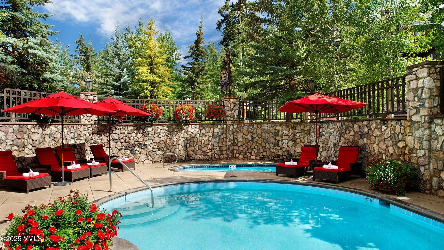 This image showcases a luxurious outdoor pool and spa area, surrounded by a stone wall and lush greenery. Red umbrellas and lounge chairs provide a comfortable and stylish relaxation space. The clear blue water of the pool invites a refreshing dip, creating an appealing and serene atmosphere.