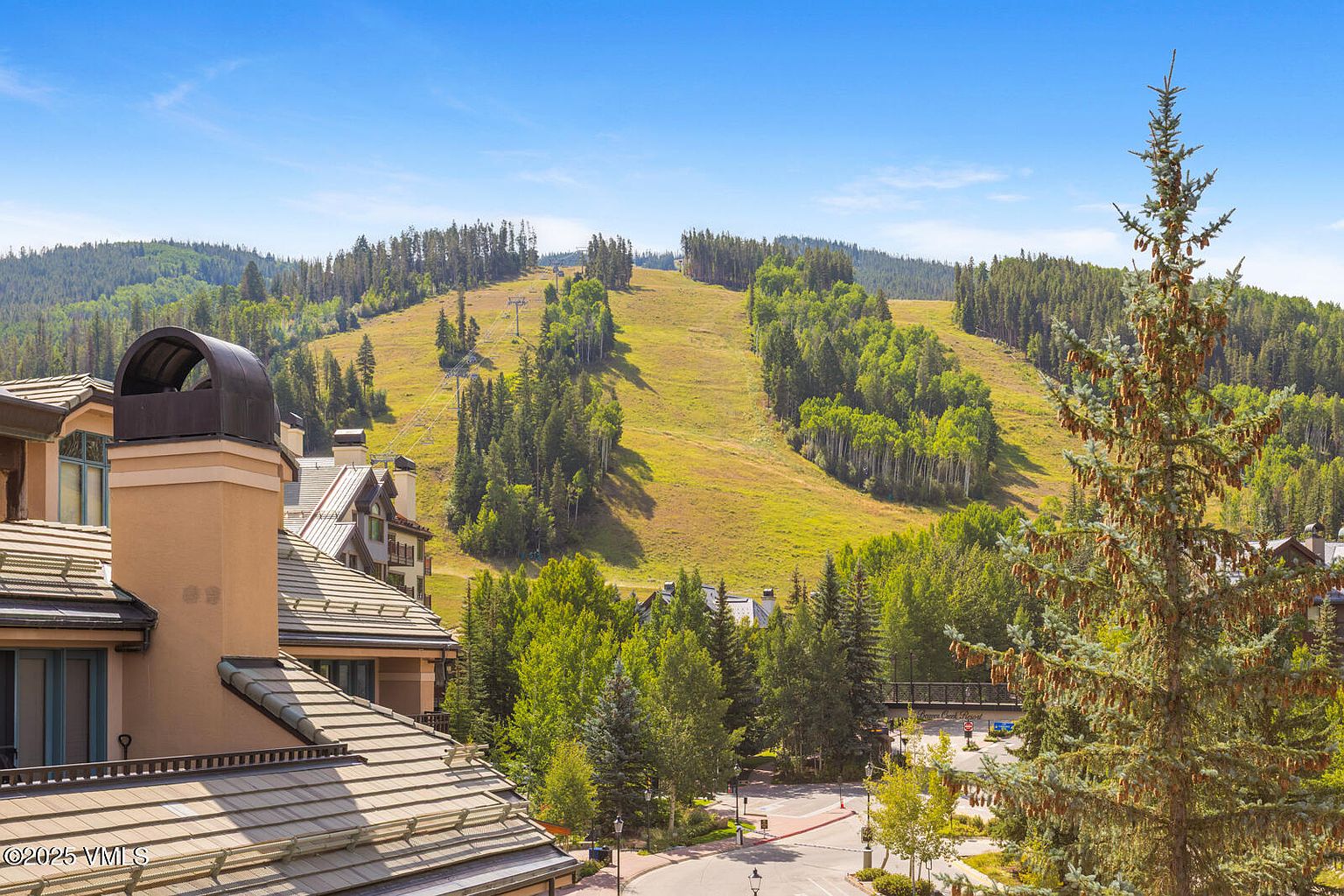 This image showcases a beautiful building complex with a mountain view. The architecture features a combination of stone and wood elements, with a distinctive chimney design. The surrounding landscape includes lush greenery and a clear blue sky, creating a serene and inviting atmosphere.