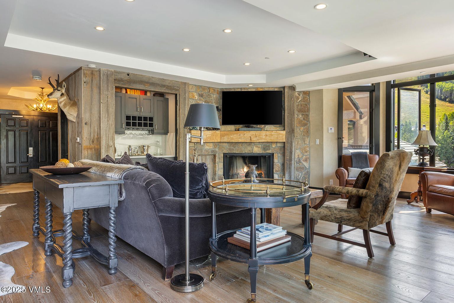This is an interior shot of a cozy living room featuring a stone fireplace with a mounted television above. The room is furnished with a comfortable dark gray sofa, a unique side table with books, and a patterned armchair. The hardwood floors and natural light from the windows create a warm and inviting atmosphere.