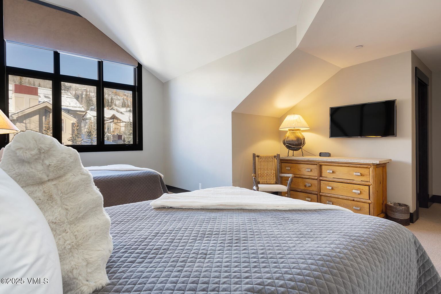 This is a cozy guest bedroom featuring two beds with gray quilted covers and white throws. A wooden dresser with a lamp and a wall-mounted TV are visible against a beige wall. A window provides natural light and a view of a snowy landscape, creating a warm and inviting atmosphere.
