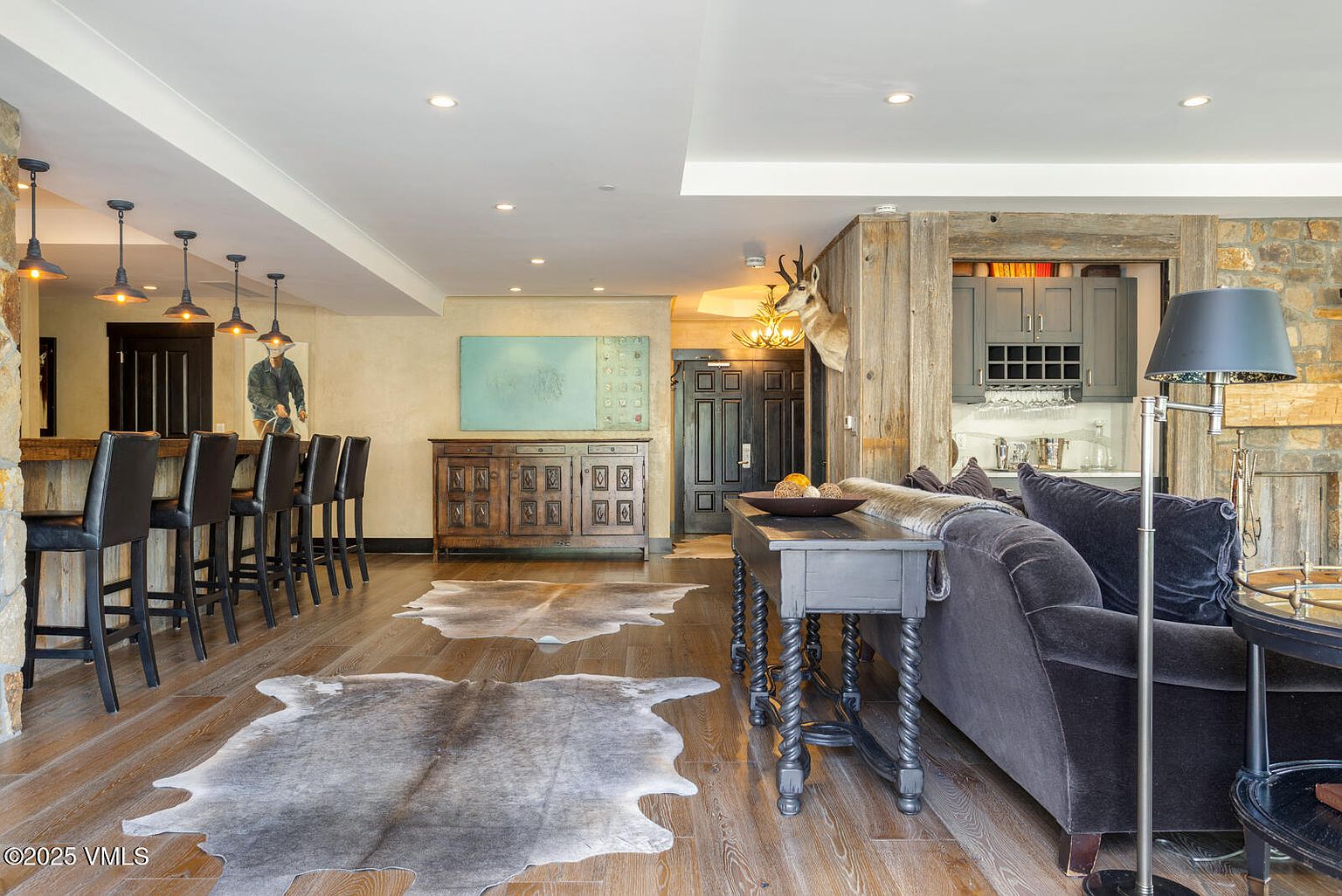 This interior shot showcases a rustic yet elegant living room. The space features hardwood floors adorned with animal hide rugs, a dark gray sofa, and a wooden console table. A bar area with stools is visible in the background, along with a stone fireplace, creating a warm and inviting atmosphere.