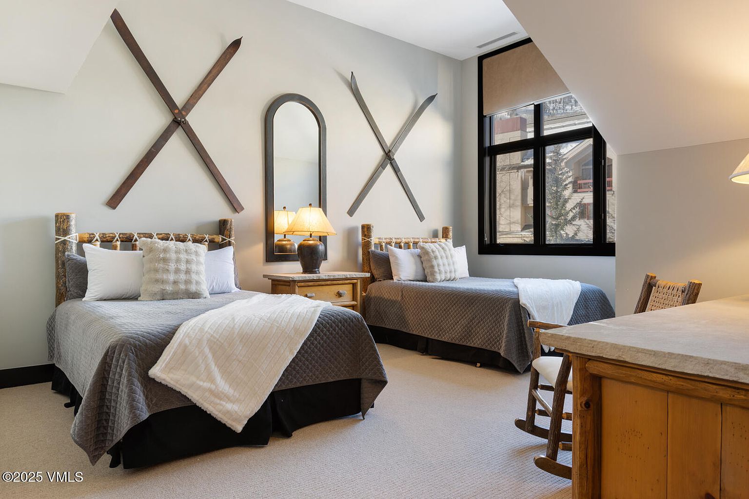 This is a cozy guest bedroom featuring two twin beds with rustic wooden frames and neutral-toned bedding. The room is decorated with vintage skis mounted on the wall and a decorative mirror, adding a touch of mountain charm. A window provides natural light, and the overall ambiance is warm and inviting.