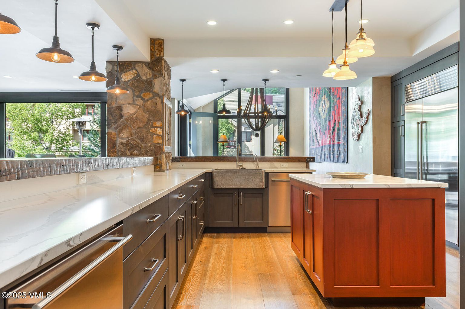 This is a well-lit kitchen featuring a long countertop with dark wood cabinets and a white marble countertop. A central island with reddish-brown cabinetry and a white marble top adds a pop of color. The kitchen also features stainless steel appliances and a large window that provides natural light and a view of the outdoors.