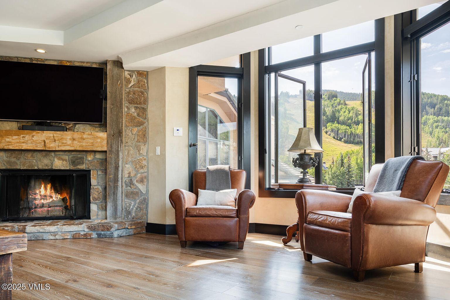 This inviting living room features a stone fireplace with a wooden mantel and a large television mounted above. Two leather armchairs are positioned near expansive windows offering views of the outside landscape. The room has a warm and comfortable ambiance, enhanced by natural light and hardwood flooring.