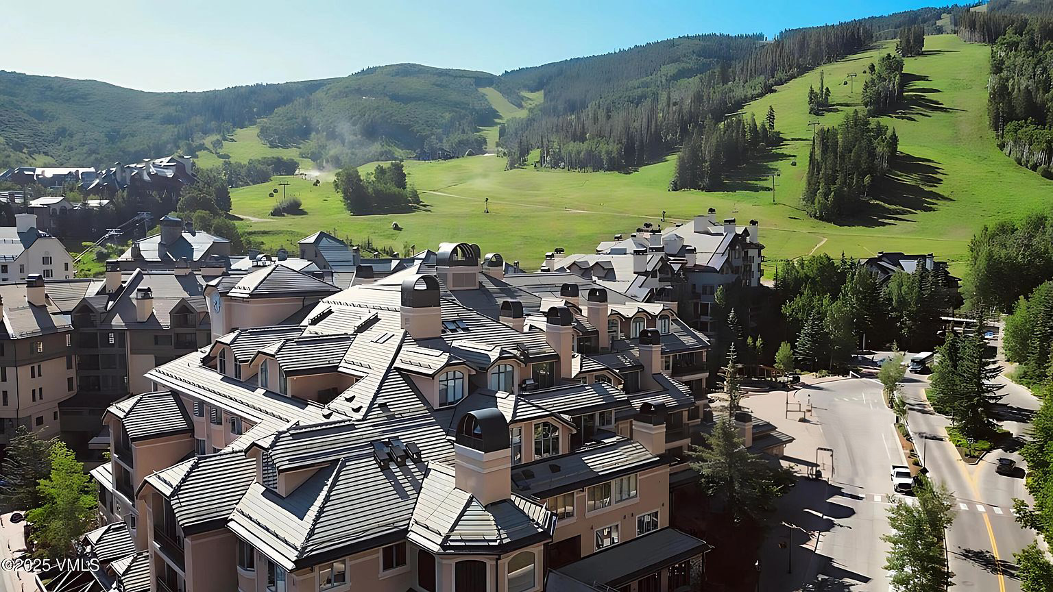 This aerial shot showcases a multi-unit residential building with a distinctive roofline featuring multiple chimneys and various roof angles. The building is surrounded by well-maintained landscaping and overlooks a scenic mountain backdrop with grassy slopes and scattered trees. The image highlights the property's proximity to natural amenities.