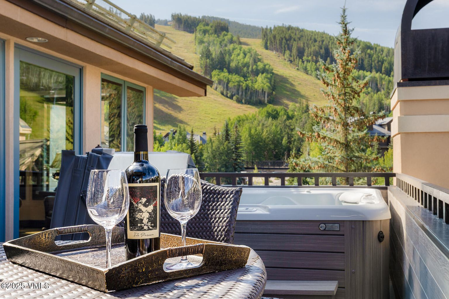 This image showcases a luxurious patio or balcony setting, complete with a hot tub and scenic mountain views. A bottle of wine and two glasses are arranged on a wicker table, suggesting relaxation and entertainment. The space is well-appointed and offers a desirable outdoor living experience.
