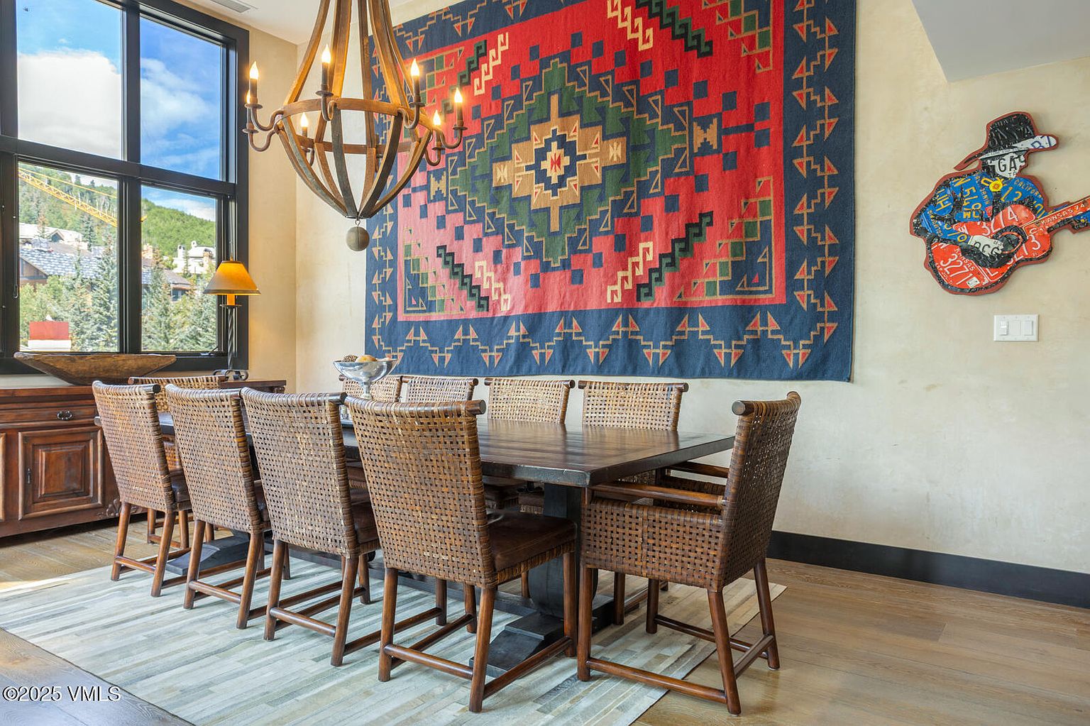 This is an interior shot of a dining room featuring a large wooden table surrounded by woven chairs. Above the table hangs a rustic chandelier, and a colorful tapestry adorns the wall, adding a touch of character. The room is well-lit by natural light streaming through a large window, creating a warm and inviting atmosphere.