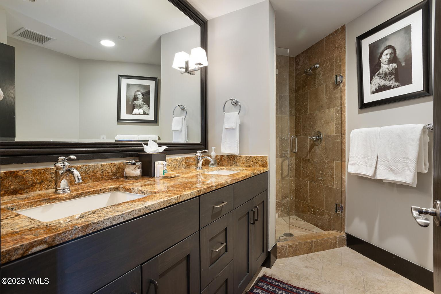 This is a well-appointed primary bathroom featuring a double vanity with a granite countertop and dark wood cabinetry. A large mirror reflects the space, including a framed artwork. A glass-enclosed shower with tiled walls is visible to the right, and the overall impression is one of luxury and functionality.