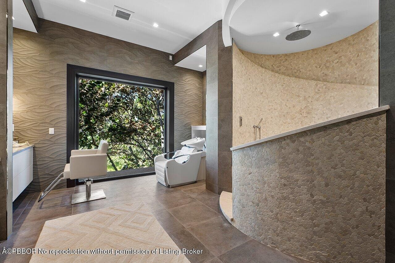 This is a luxurious primary bathroom featuring a modern design with a unique shower area adorned with pebble-like tiles and a rainfall showerhead. The room includes a salon-style chair and sink, positioned near a large window that offers natural light and a view of the greenery outside. The flooring is tiled, and the walls have a textured, wave-like pattern, contributing to the spa-like atmosphere.