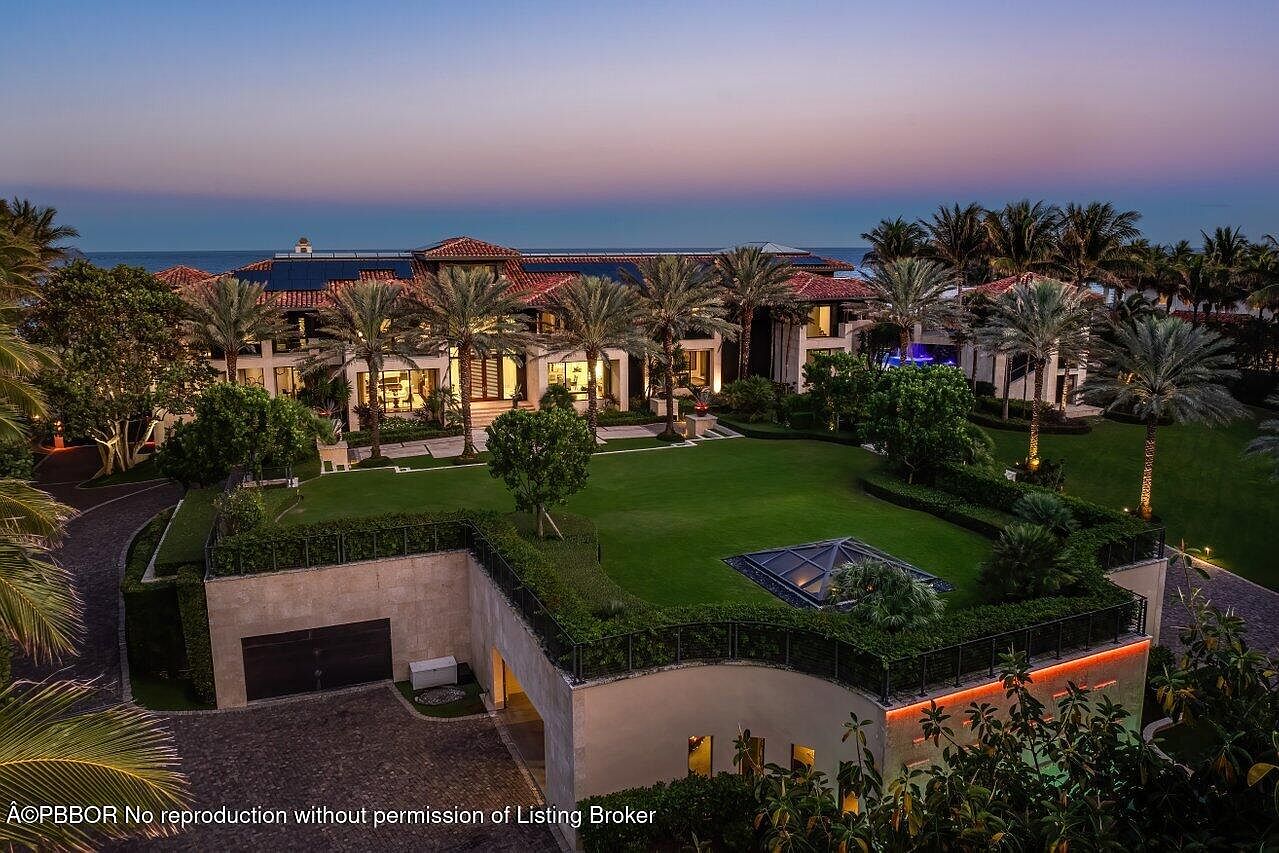 This aerial view showcases a luxurious estate with a red tile roof, lush green lawns, and numerous palm trees. The property features a unique underground garage entrance topped with greenery, adding to its sophisticated design. The ocean is visible in the background, suggesting a prime beachfront location.