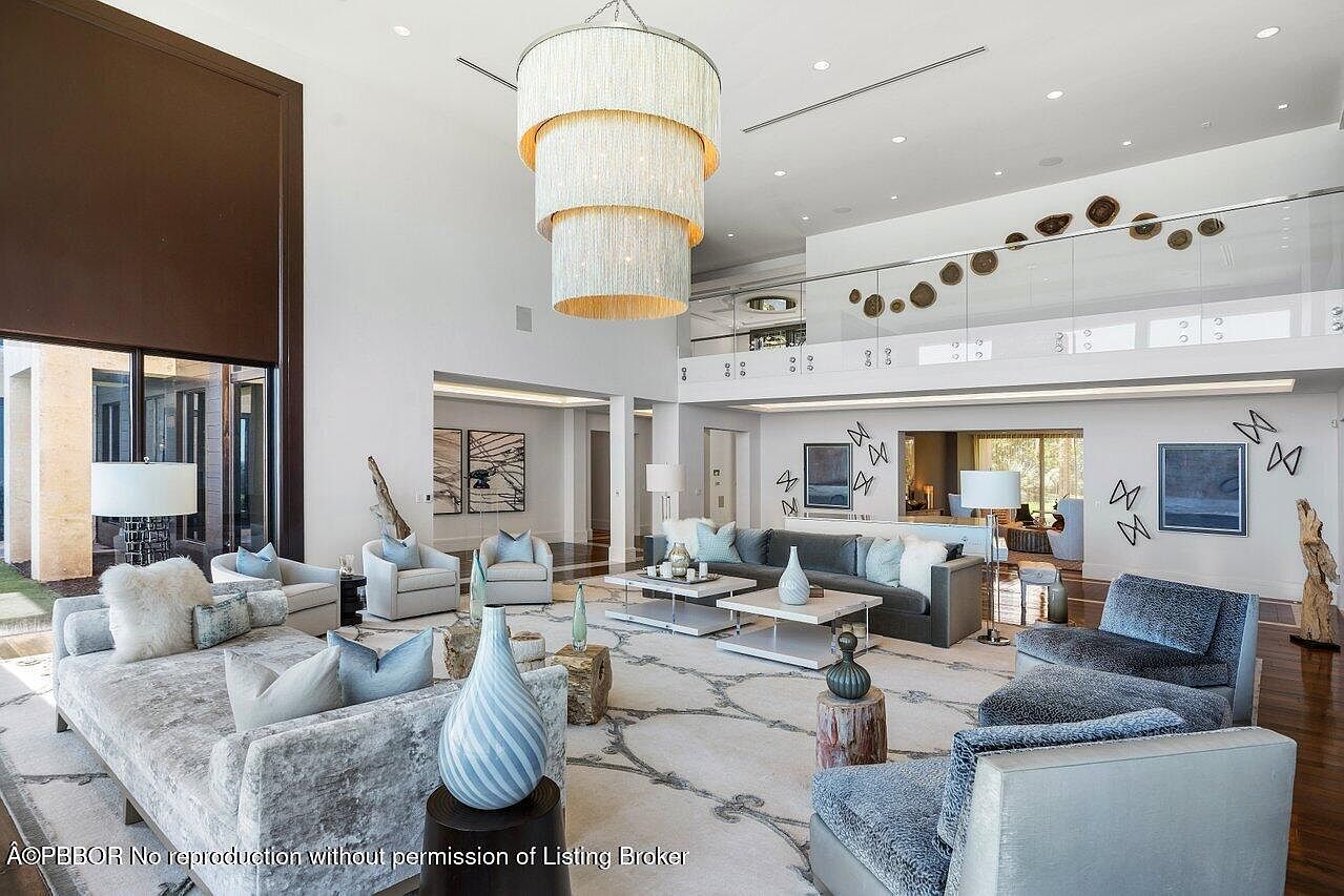 This is a spacious and luxurious living room featuring high ceilings and an open floor plan. The room is furnished with plush sofas, armchairs, and stylish coffee tables, all arranged on a large patterned rug. A grand chandelier hangs from the ceiling, adding to the room's elegant ambiance.
