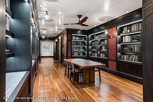 This is an interior shot of a sophisticated home office or study. The room features dark wood built-in bookshelves filled with books, a large wooden table serving as a desk, and hardwood floors. The lighting is warm and inviting, creating a comfortable and professional atmosphere.