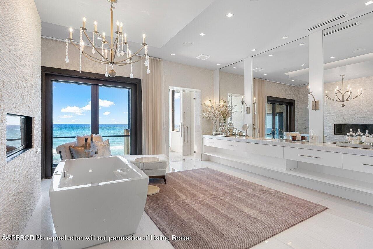 This is a luxurious primary bathroom featuring a freestanding bathtub with ocean views through a large window. The room is decorated with a modern chandelier, a long vanity with mirrors, and a neutral-toned rug. The overall impression is one of opulence and relaxation.