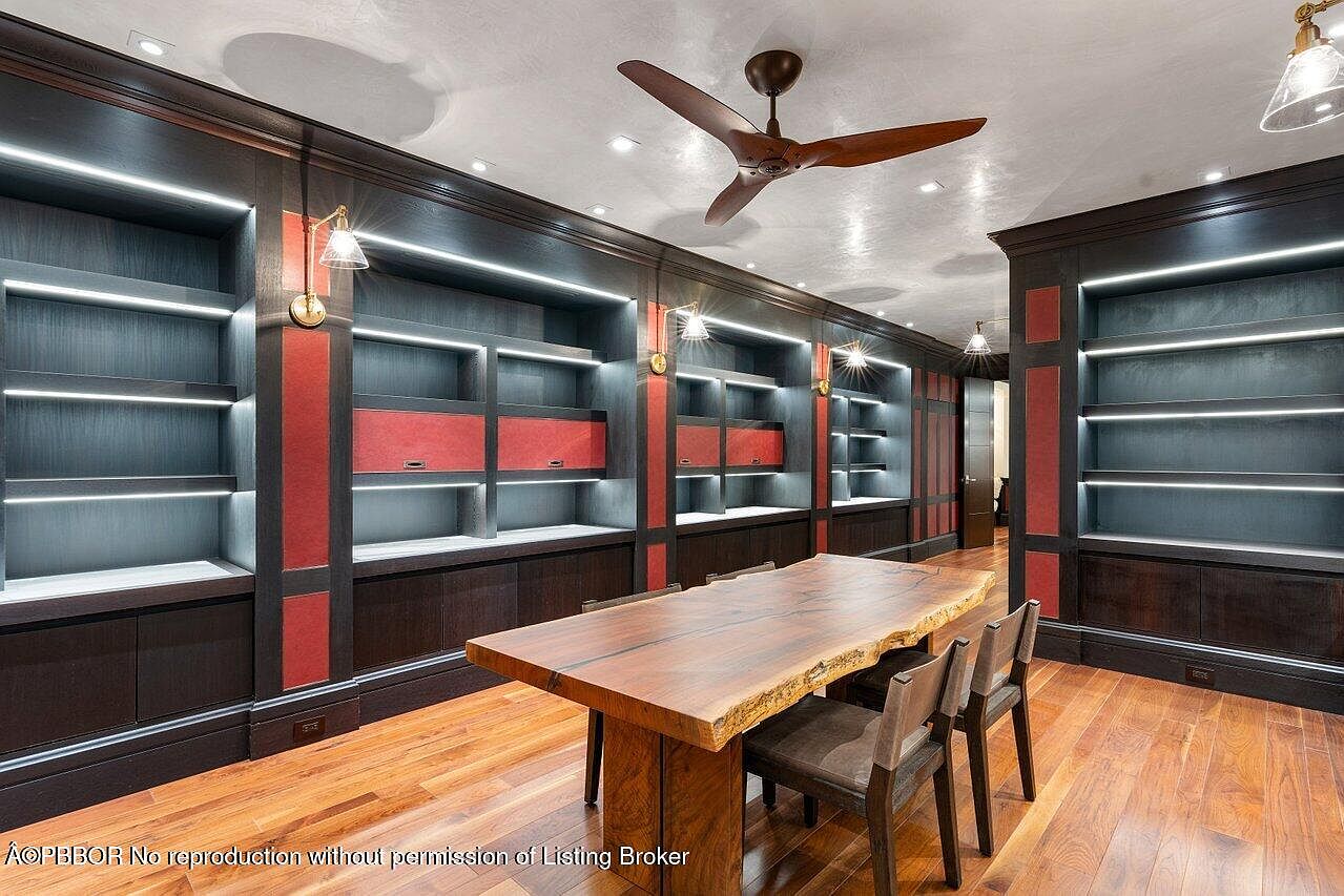 This is an interior shot of a home office or study, featuring custom built-in bookshelves with integrated lighting. A large, natural-edge wooden table with chairs sits in the center of the room, creating a functional workspace. The hardwood flooring and ceiling fan add to the room's character and appeal.