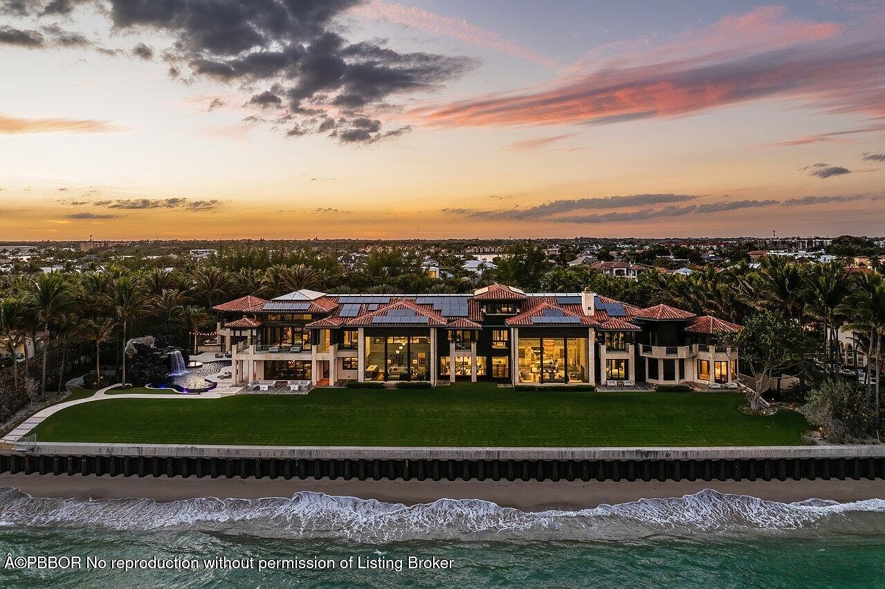 This aerial view showcases a luxurious beachfront estate at sunset. The large home features a red tile roof, multiple windows, and a sprawling green lawn leading to the beach. The ocean waves gently lap against the shore, and the sky is painted with vibrant colors.