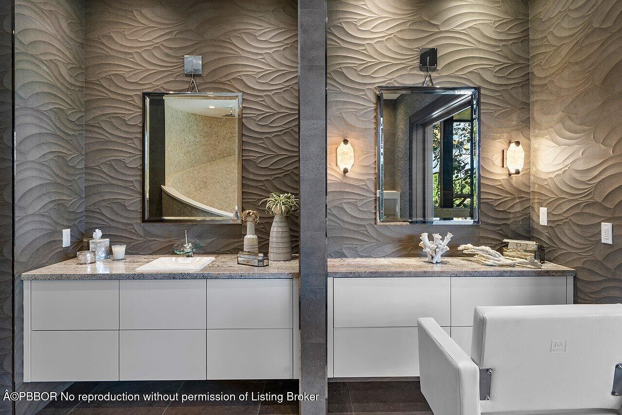 This is a primary bathroom featuring dual vanities with white cabinets and granite countertops. The walls are adorned with textured, wave-patterned tiles, and each vanity has a rectangular mirror with modern lighting fixtures. The overall design is contemporary and luxurious, creating a spa-like atmosphere.