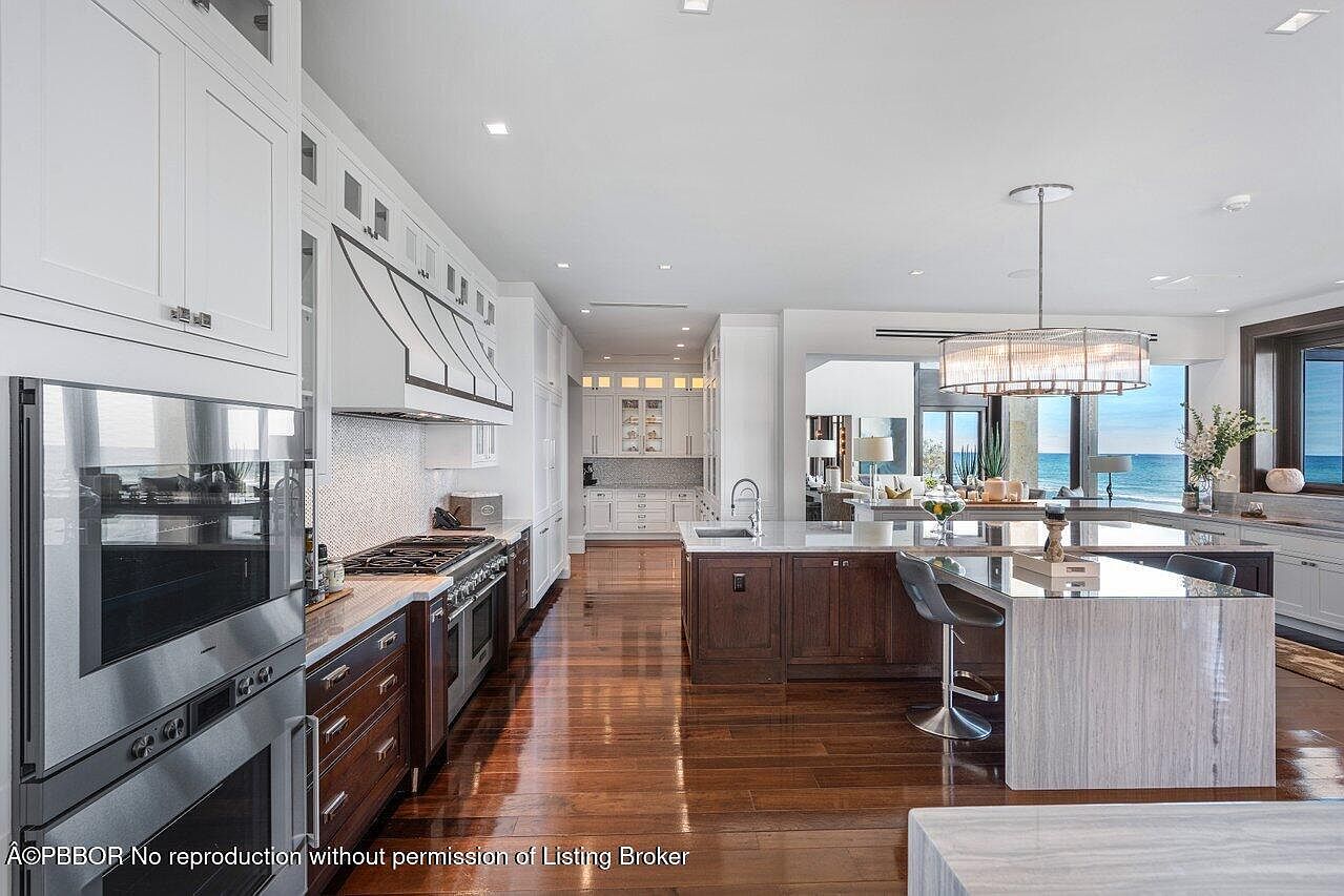 This is a bright and spacious kitchen featuring dark wood floors, white cabinetry, and stainless steel appliances. A large island with seating provides a central gathering point, and the open layout connects to a living area with ocean views. The kitchen is well-lit with recessed lighting and a decorative chandelier, creating an inviting and luxurious atmosphere.