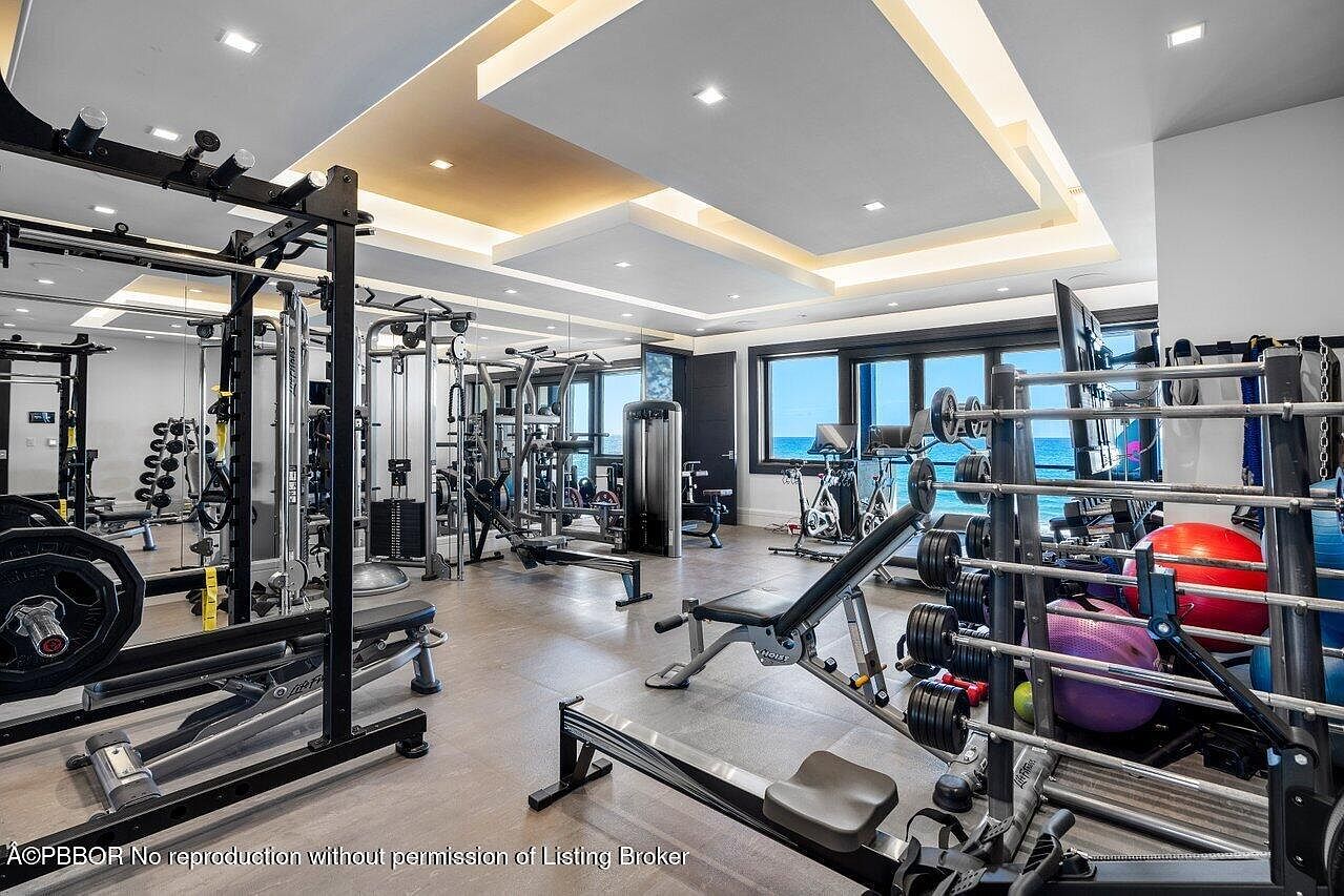 This is a spacious home gym featuring a variety of exercise equipment, including weight racks, benches, and cardio machines. Large windows offer a view of the ocean, creating an inspiring workout environment. The room is well-lit with modern ceiling fixtures and has a clean, organized appearance.