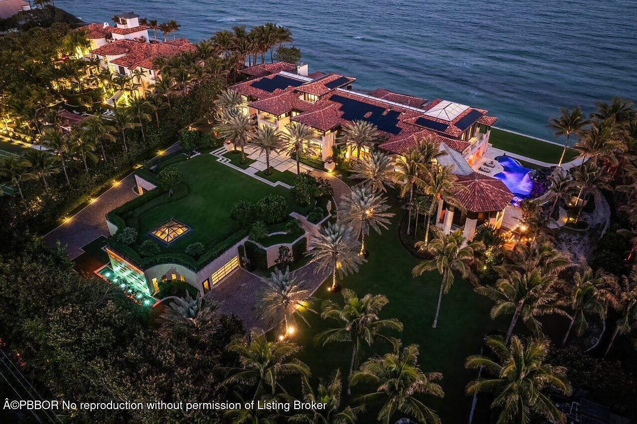 This aerial view showcases a luxurious estate with a red tile roof, lush landscaping, and a stunning oceanfront location. The property features a large main house, a separate guest house, a swimming pool, and meticulously manicured lawns adorned with palm trees. The overall impression is one of opulence and exclusivity.