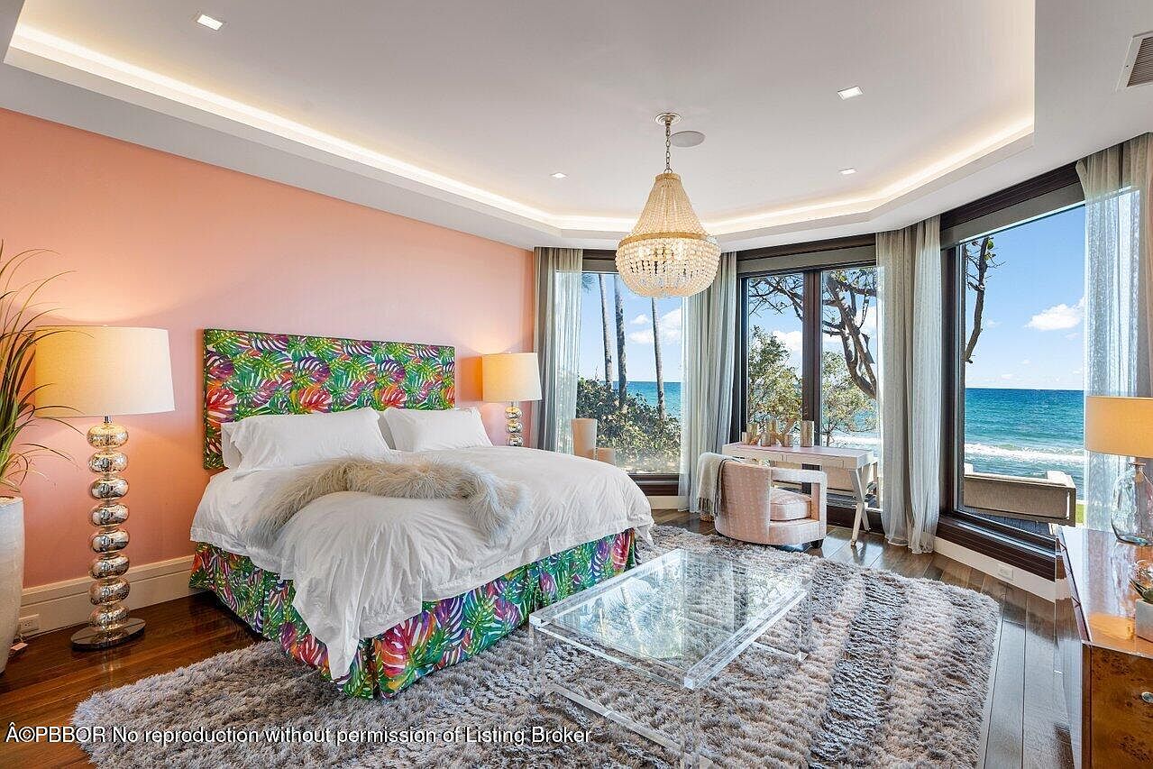 This primary bedroom features a vibrant tropical-themed headboard and bed skirt, complemented by crisp white linens and a plush throw. A crystal chandelier hangs from the ceiling, and large windows offer stunning ocean views, creating a luxurious and serene atmosphere. The room is furnished with a glass coffee table and a small desk area, adding functionality to the space.
