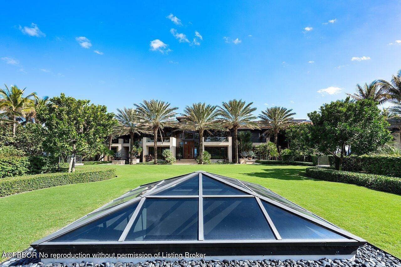 This image showcases a meticulously maintained yard and garden, featuring lush green grass, manicured hedges, and mature palm trees. A unique glass pyramid structure sits in the foreground, adding an architectural element. The property's exterior exudes luxury and tranquility, suggesting a high-end residential estate.