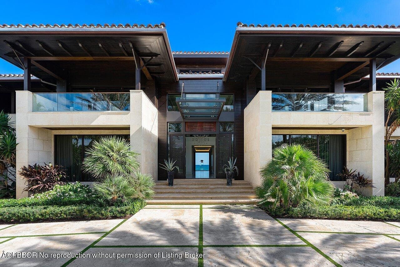 This is a front view of a luxurious home's entryway, featuring a modern architectural design with a combination of light stone and dark wood elements. The entrance is elevated with steps leading to a glass-covered doorway, flanked by manicured greenery and palm trees. The driveway is paved with large stone tiles separated by strips of grass, adding to the property's curb appeal.