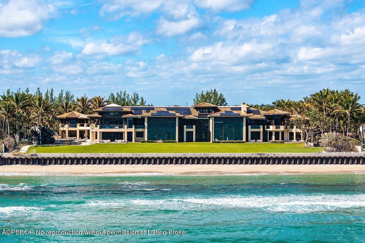 This image showcases the rear exterior of a luxurious beachfront property. The architecture features large windows, a tile roof, and a well-manicured lawn extending to the beach. The property is bordered by palm trees and the ocean, creating a serene and upscale coastal living environment.