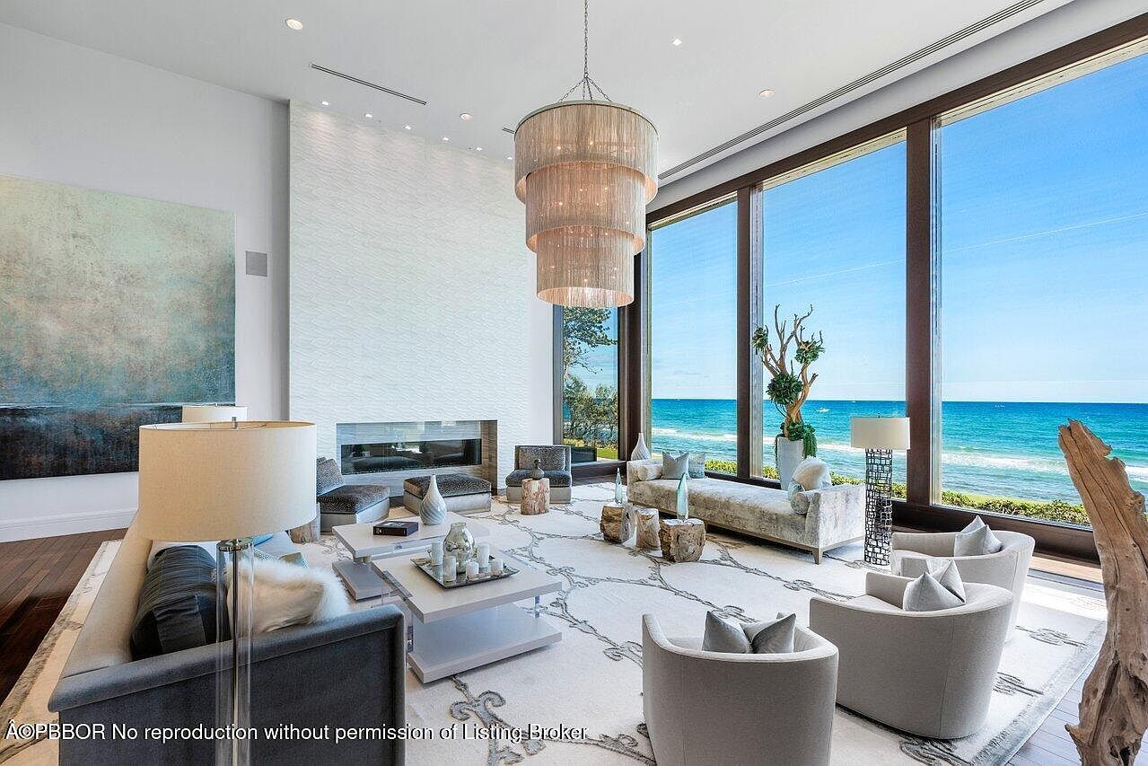 This is a luxurious living room with floor-to-ceiling windows offering stunning ocean views. The room features a modern fireplace, a tiered chandelier, and stylish furniture arranged on a patterned rug. The overall impression is one of elegance and tranquility, perfect for high-end living.