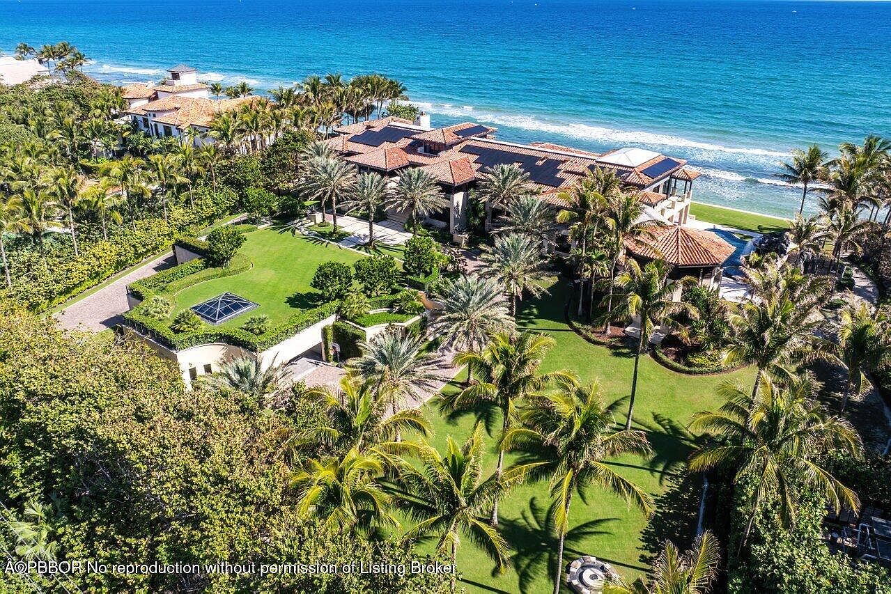 This aerial shot showcases a luxurious beachfront estate with meticulously manicured lawns, lush palm trees, and a sprawling main residence featuring a terracotta tile roof. The property extends to the oceanfront, offering stunning views of the turquoise water and a private beach area. The overall impression is one of opulence and serene coastal living.